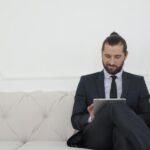 Un homme d'affaires en costume assis sur un canapé utilisant une tablette, souriant.
