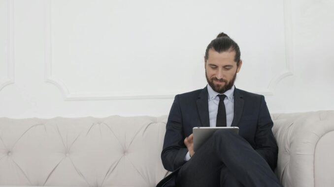 entrepreneur Un homme d'affaires en costume assis sur un canapé utilisant une tablette, souriant.